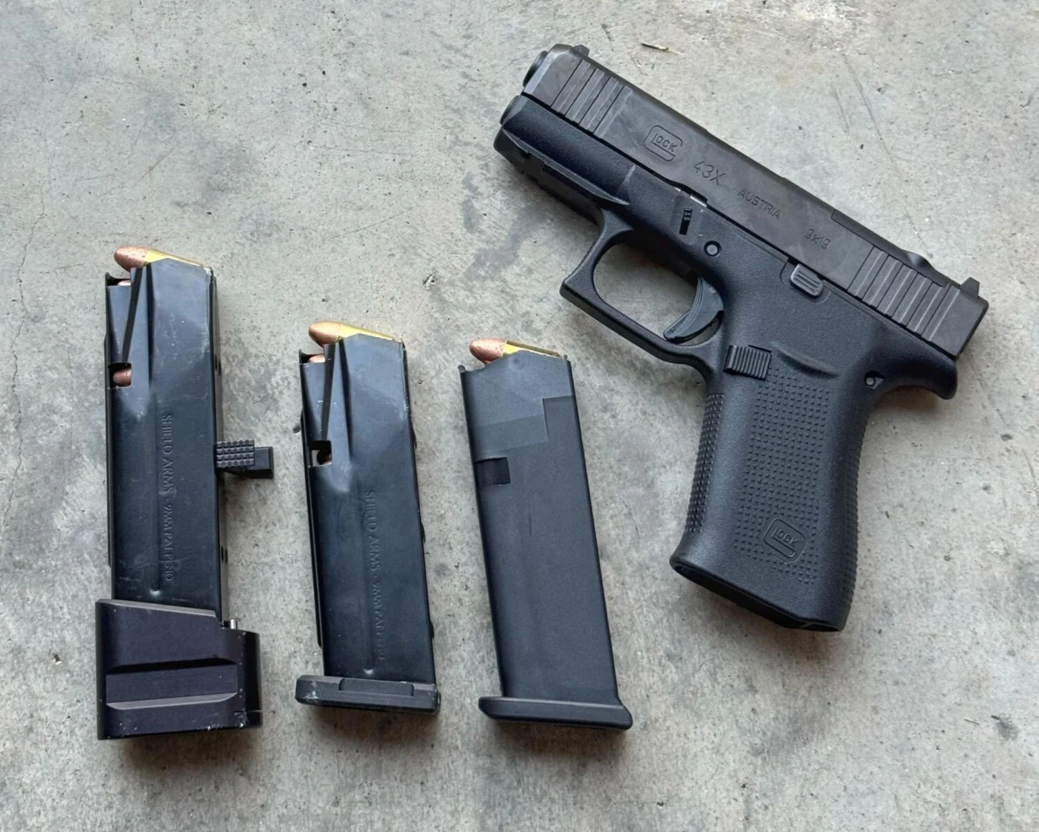 A Glock 43X pistol next to two Shield Arms steel magazines, one fitted with an extension, and a factory Glock magazine