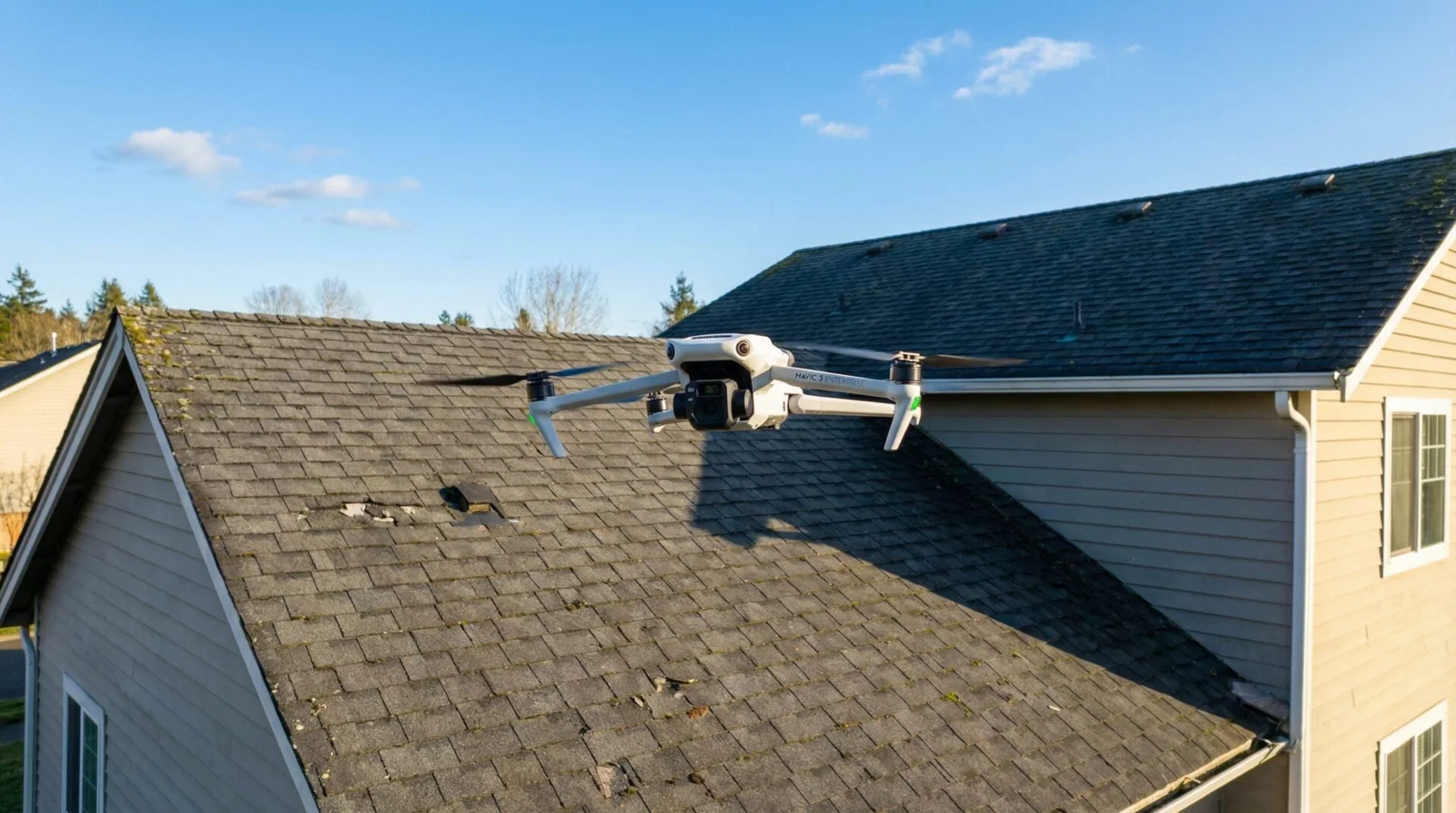 DJI Mavic 3 Enterprise quadcopter hovering a few feet above the shingled roof of a suburban two-story house on a clear day