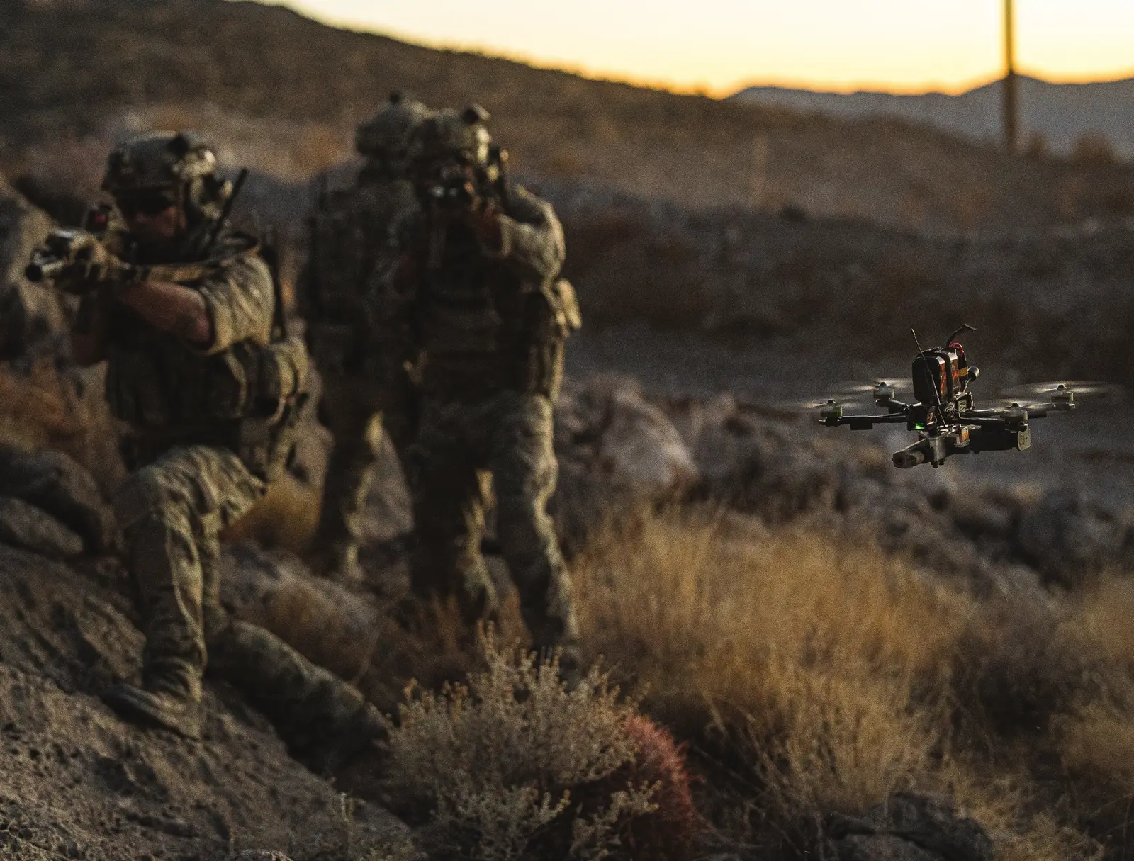 Soldiers in camouflage aiming rifles across a rocky desert slope while a small quadcopter hovers beside them at dusk