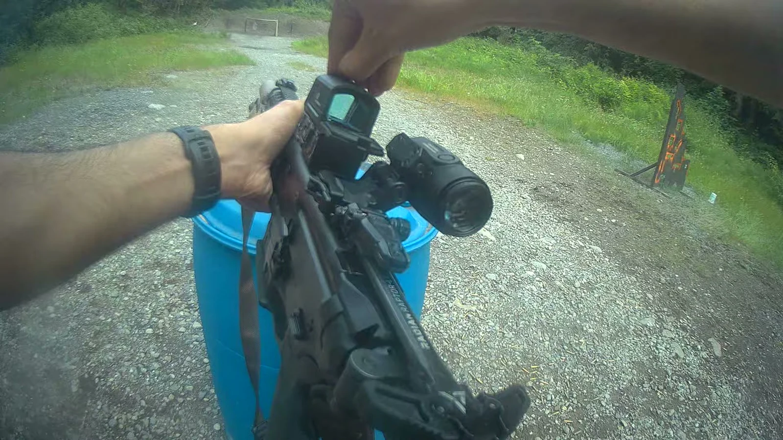 Chest camera frame showing a hand reaching over the top of the ROMEO8T-AMR housing to open the front flip cap, with the magnifier beside the sight and wet gravel underfoot