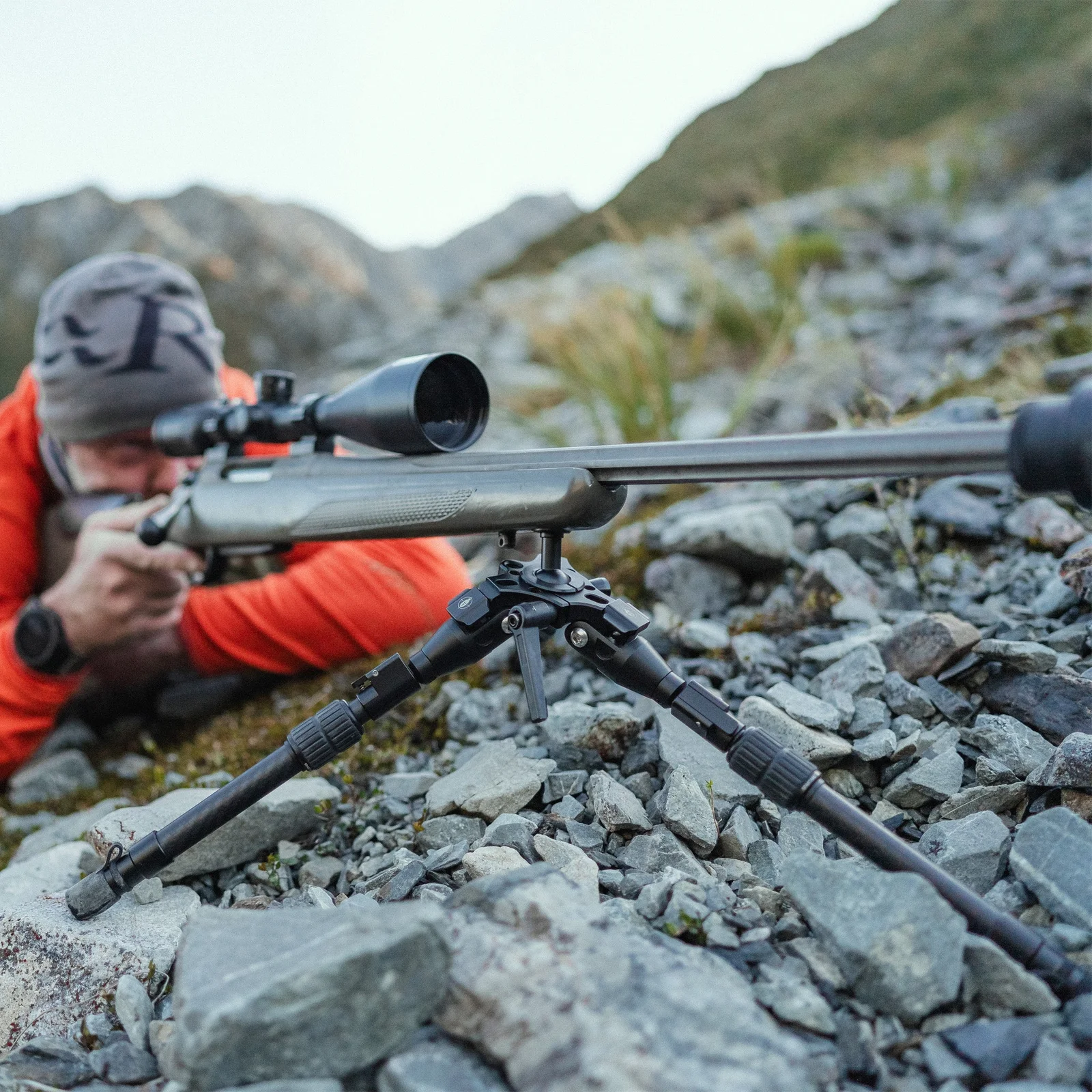 Precision bolt-action hunting rifle supported by a bipod on a rocky mountain slope with a hunter aiming in the background