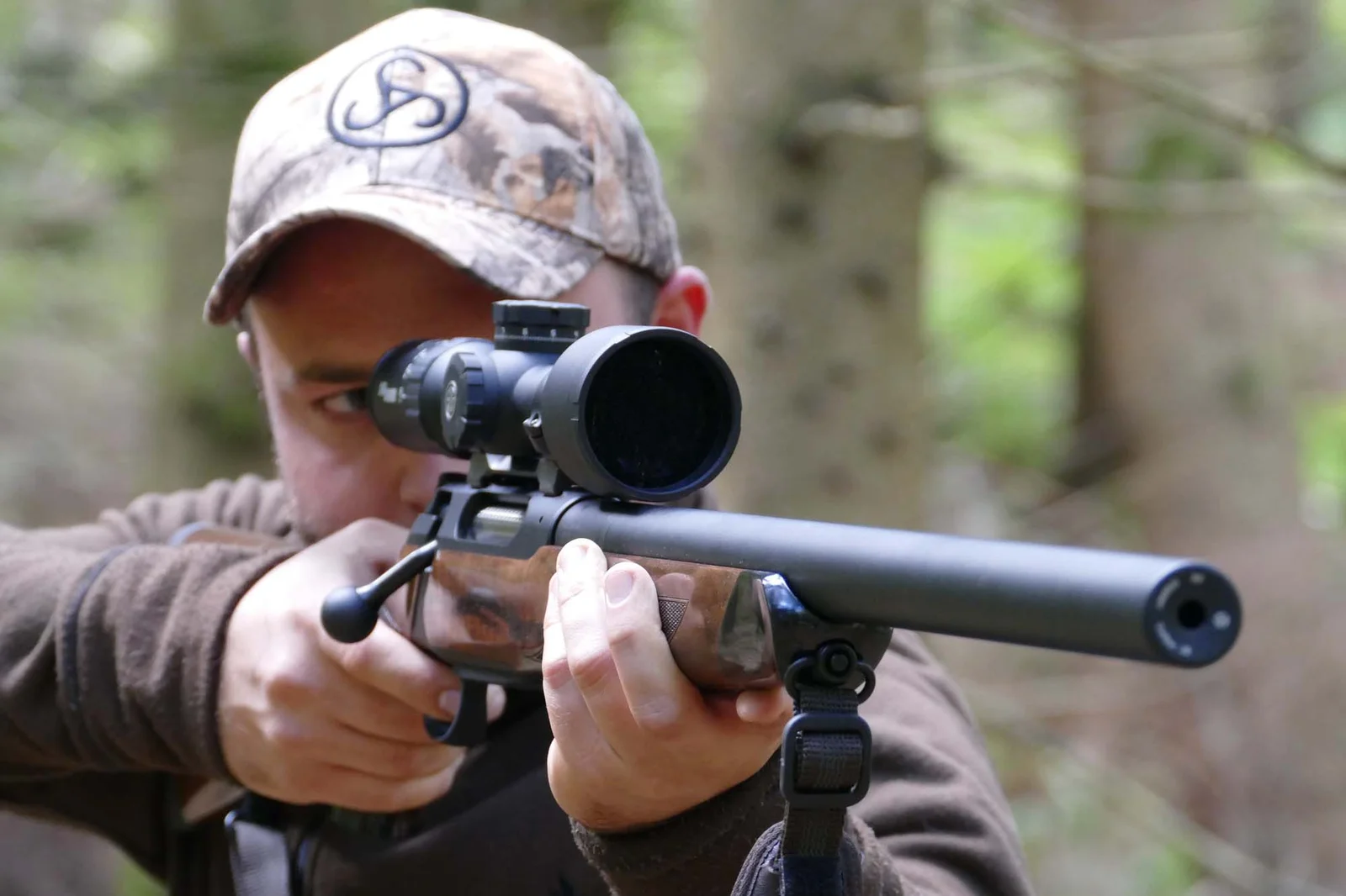 Hunter in a camouflage cap shouldering a scoped bolt-action rifle with a suppressor mounted, in a forest
