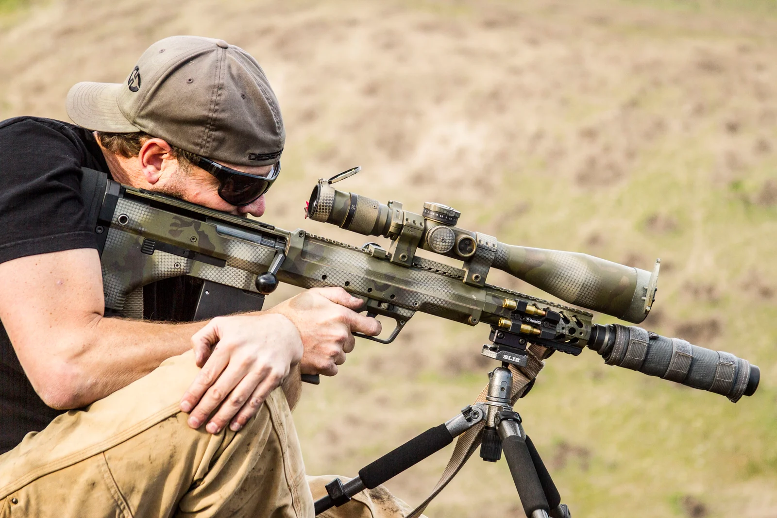 Shooter kneeling behind a camouflage-painted bolt-action rifle with a scope and a suppressor, resting on a tripod at an outdoor range