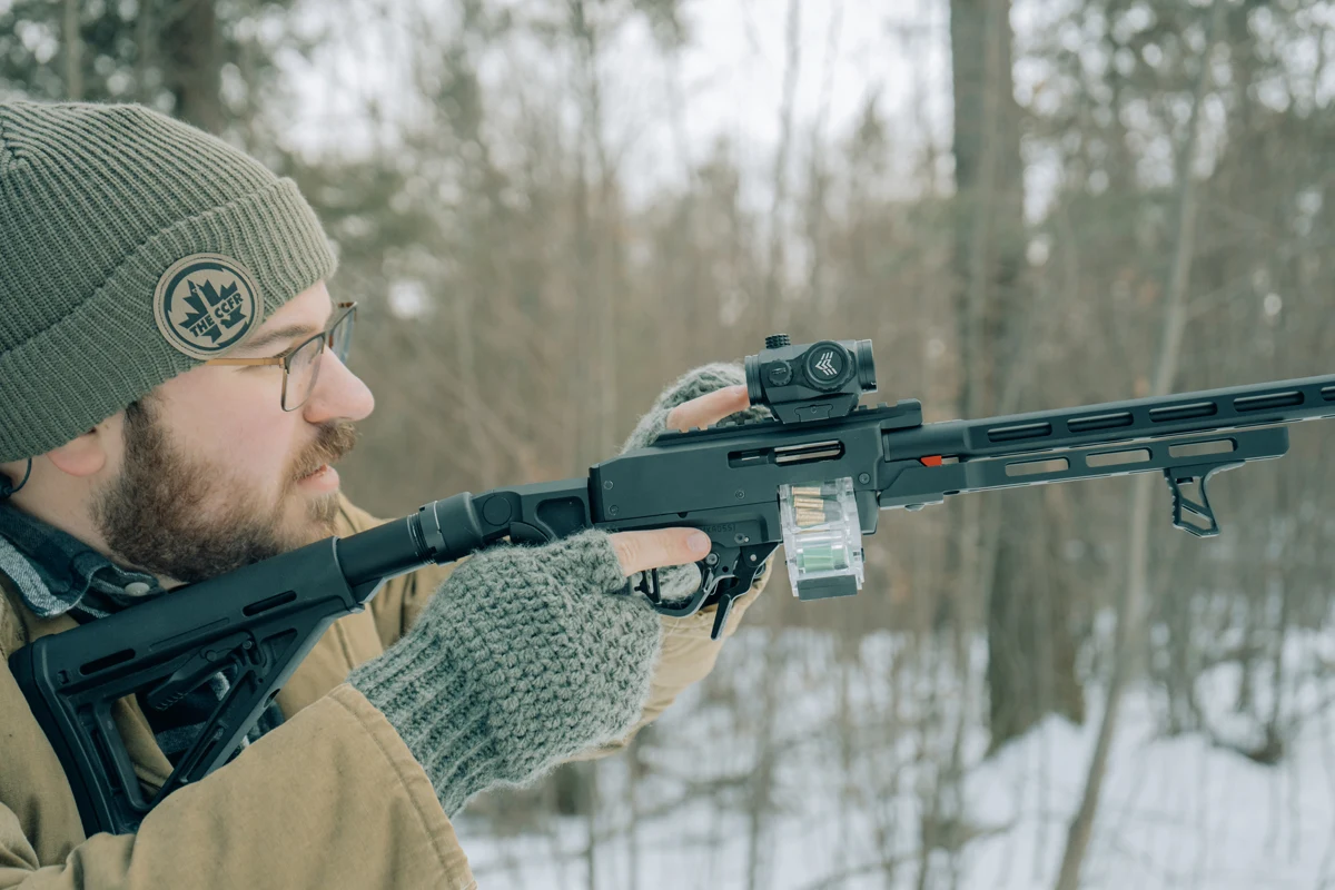 Shooter in a winter forest shouldering a TANDEMKROSS ADX-receiver rifle with a red dot, M-LOK barrel sleeve, and clear magazine