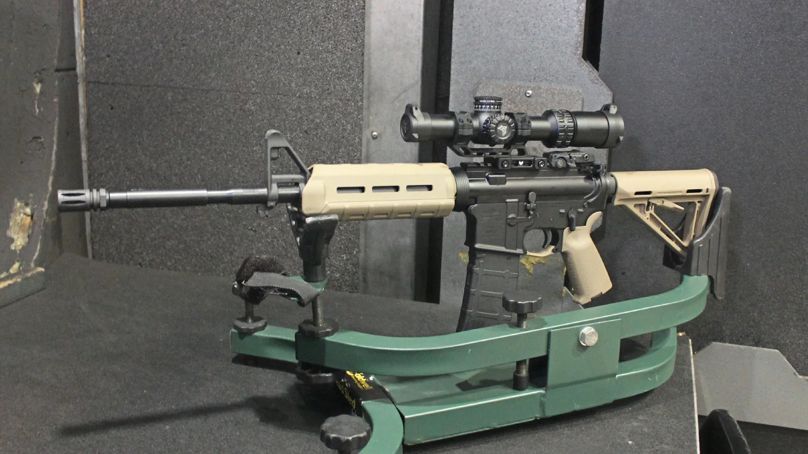 A semi-automatic AR-15 with tan Magpul furniture and a low-power variable optic resting on a green shooting rest at an indoor range
