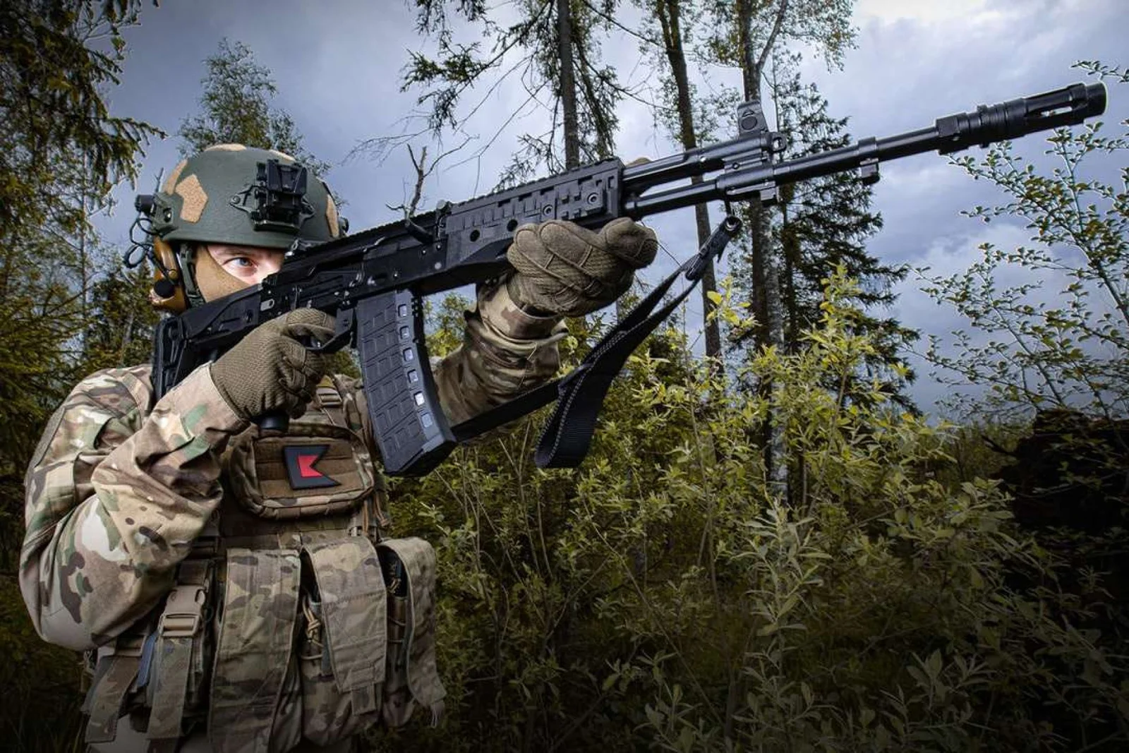 A soldier shoulders an AK-12 rifle with side-folding stock, full-length top rail, and a polymer 30-round magazine in a wooded setting
