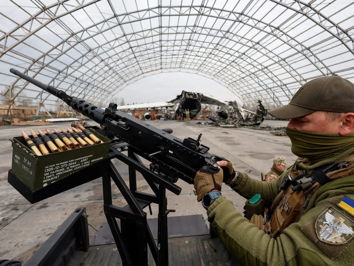 A Ukrainian soldier mans a pintle-mounted Browning M2 heavy machine gun with a linked belt in a green ammunition can, with a destroyed large aircraft visible behind him