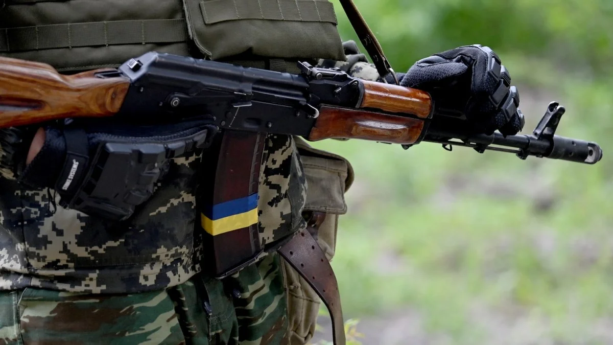 Close view of a Ukrainian soldier carrying an AK-74 pattern rifle with wood furniture, a plum polymer magazine marked with a Ukrainian flag, and a plain plate carrier
