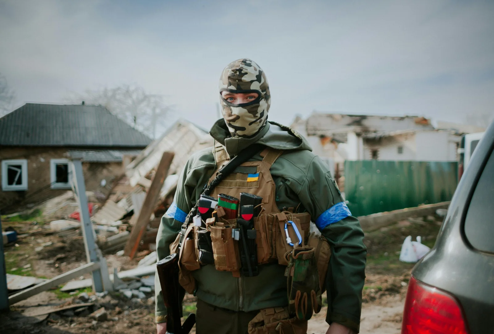 Ukrainian soldier in a tan plate carrier with AK magazines in front pouches, blue identification armbands, standing in front of a destroyed house