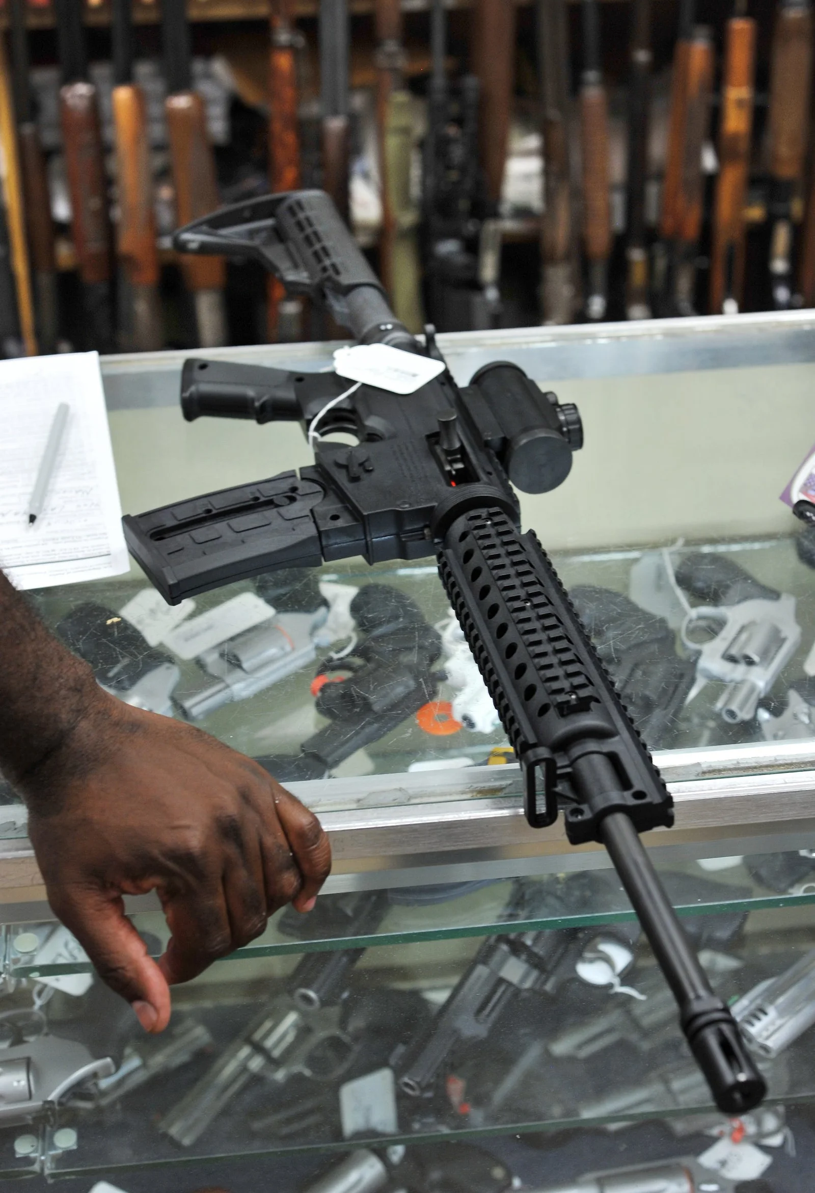 A black AR-15 with a quad-rail handguard, collapsible stock, pistol grip, red dot optic, and 30-round magazine held over a gun store display case