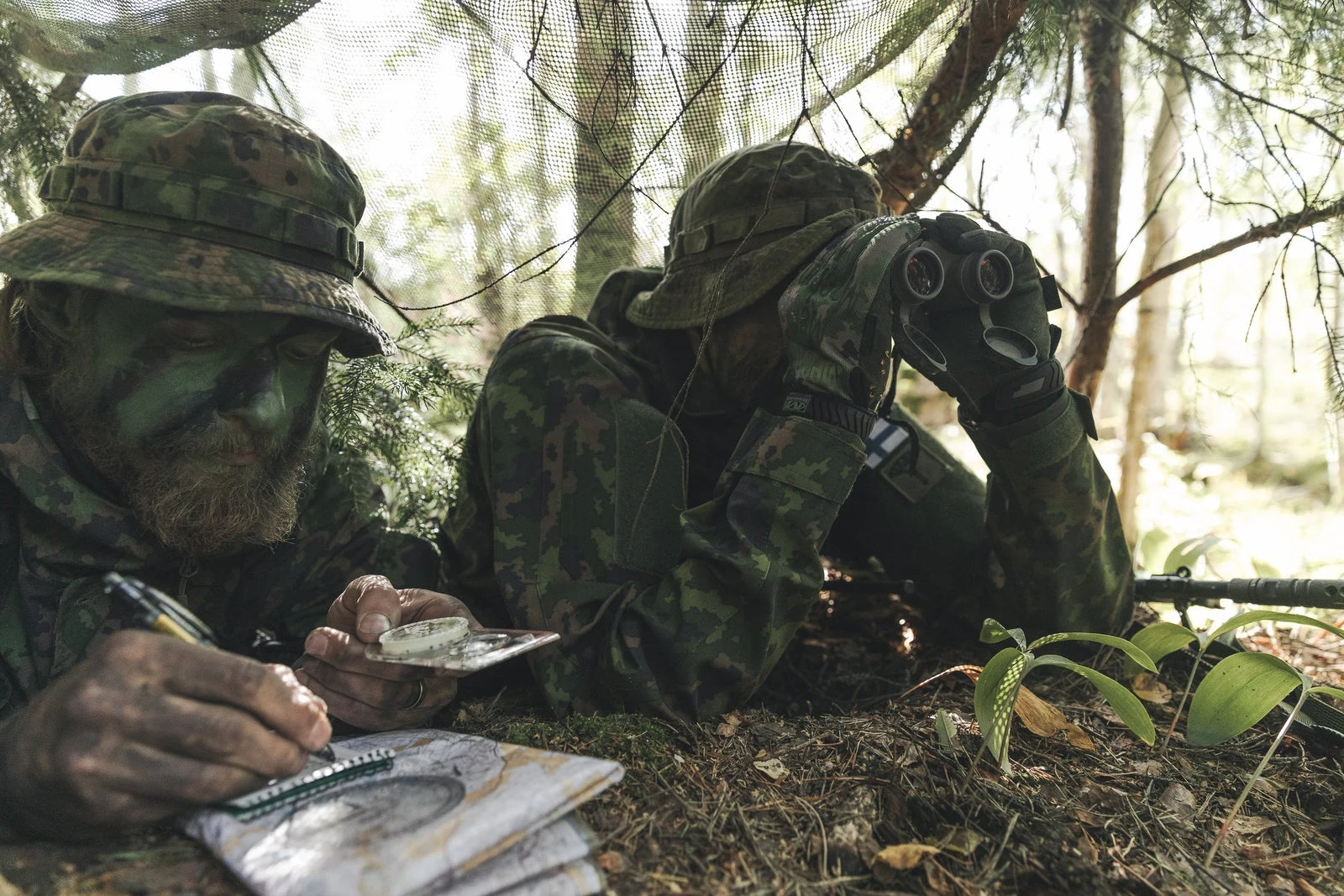 Two soldiers in Finnish M05 camouflage under a camouflage net in a forest, one glassing with binoculars and one plotting a position on a map with a compass