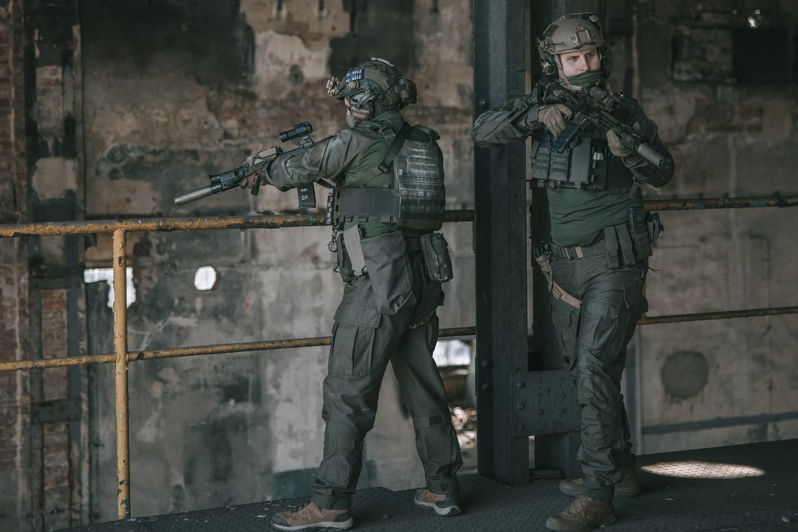 Two operators in Varusteleka combat shirts and field trousers with plate carriers, helmets, and suppressed rifles moving through a derelict industrial building