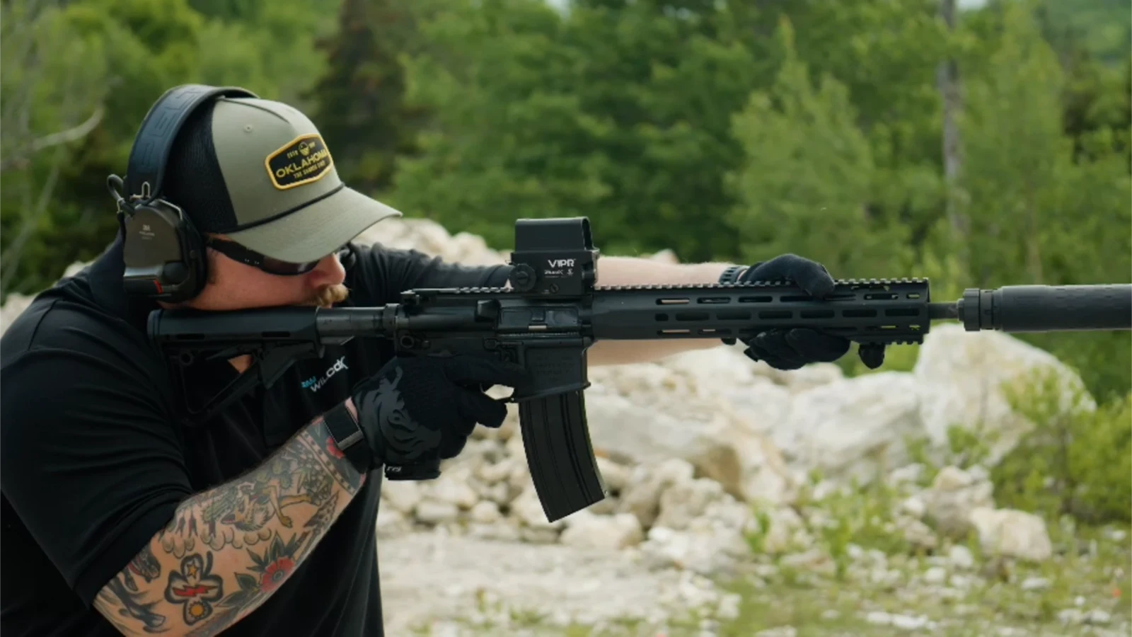 Shooter in a cap and electronic hearing protection firing a suppressed AR-15 carbine topped with a Wilcox VIPR sight on an outdoor range
