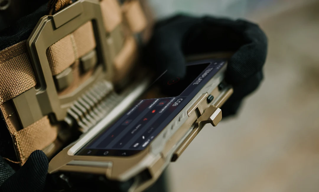 Gloved hands holding a rugged phone in a chest rig mount displaying the VIPR app CQB reticle template screen