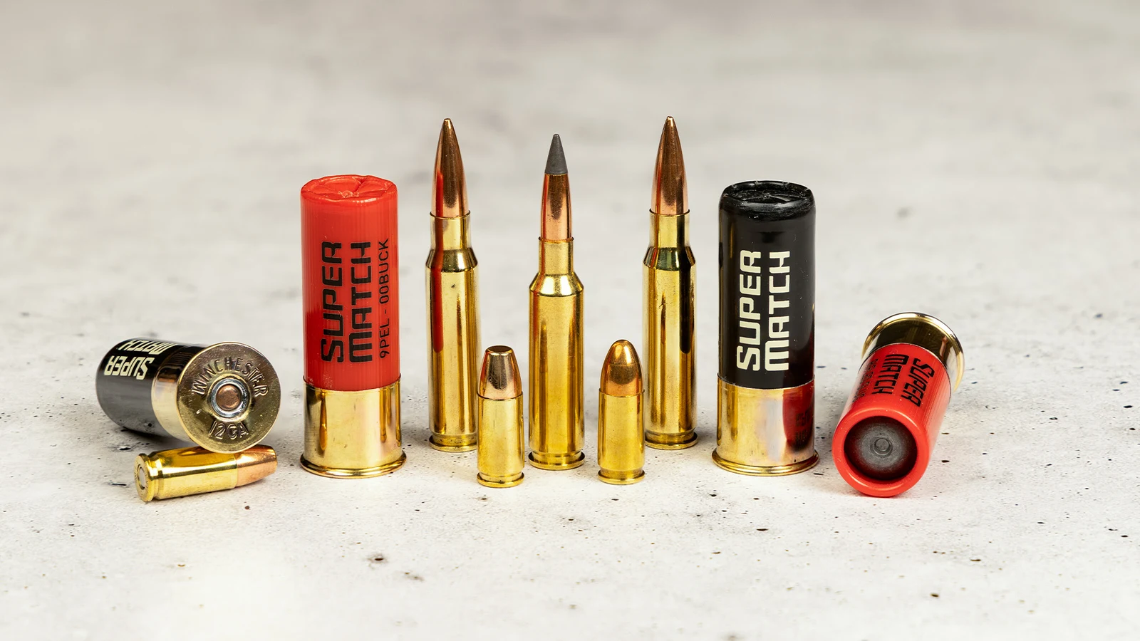 Loose Winchester Super Match cartridges including 12 gauge 00 buck, rifled slug, rifle rounds, and 9mm standing on concrete