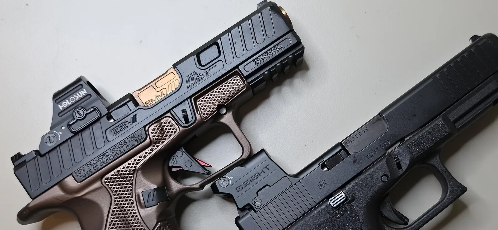 A ZEV Technologies Glock slide with lightening cuts and a Holosun red dot mounted, fitted to a bronze stippled frame with a gold barrel, beside a black Glock slide carrying an enclosed OSight red dot