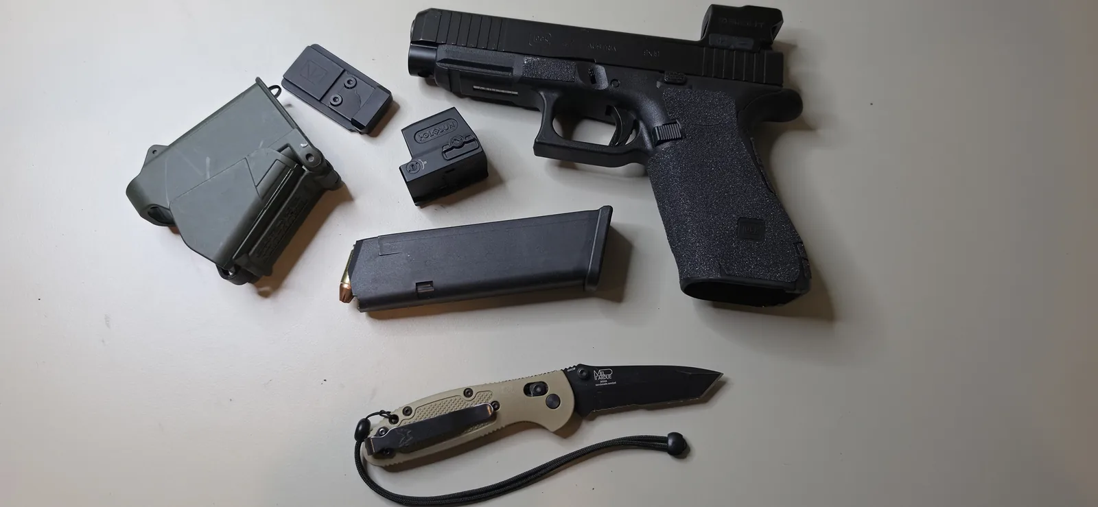 Full-size Glock MOS pistol with an enclosed red dot mounted, staged beside an MOS adapter plate, a second red dot, a magazine, and a folding knife