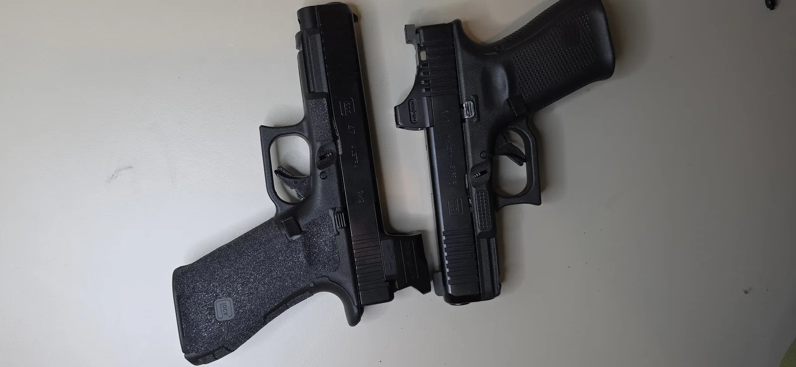 A full-size Glock 47 and a compact Glock 19 Gen5 laid side by side with their grips toward the viewer, showing how much further the full-size grip extends below the trigger guard