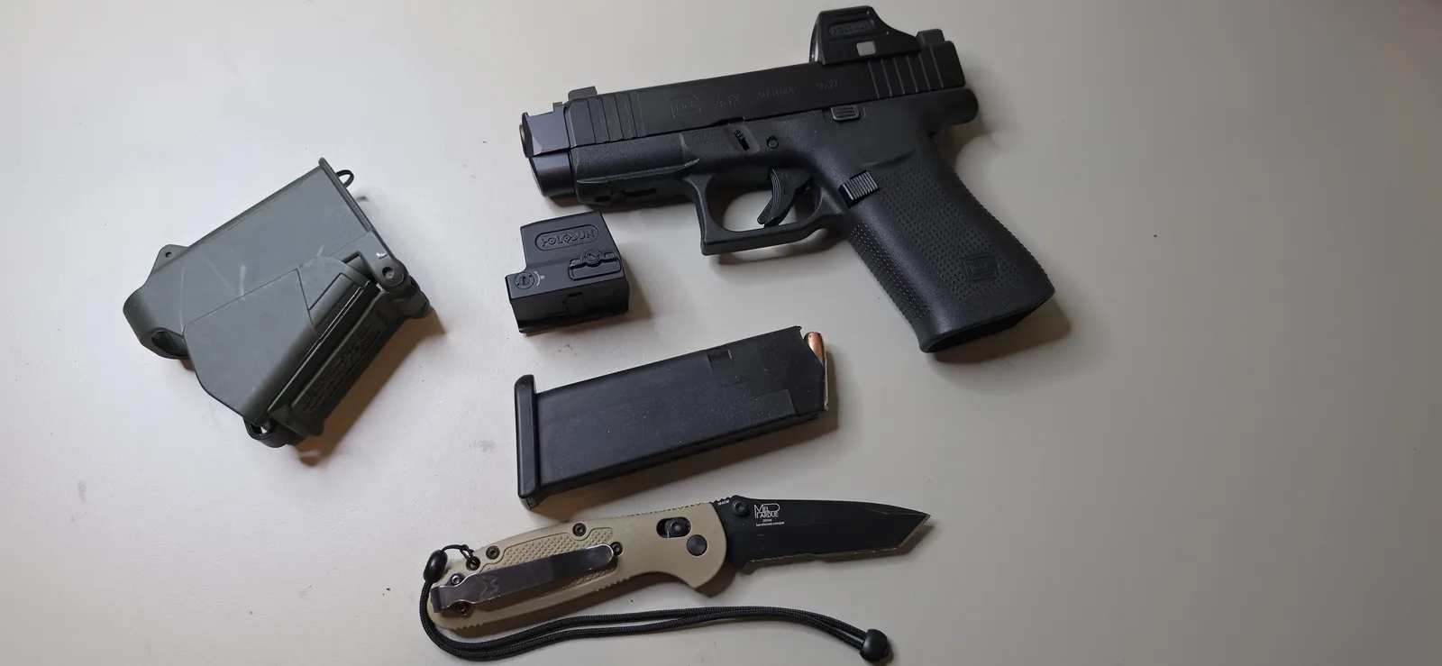 Glock 43X running a Radian Ramjet barrel and Afterburner compensator with a Holosun red dot mounted to the slide, laid out beside a spare optic, a magazine, a polymer magazine carrier, and a folding knife