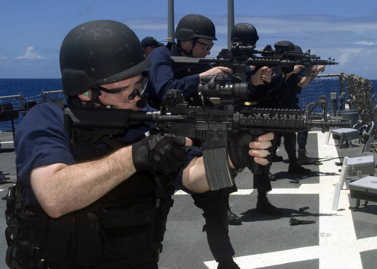 Sailors firing MK18 Mod 0 carbines on a ship deck; the foreground rifle shows a fixed triangular front sight base with a two-piece drop-in quad rail clamped around it, an aluminum GI magazine, and an A2 pistol grip