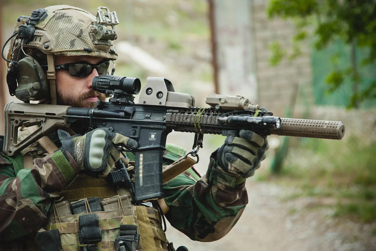 Operator shouldering a short 5.56 carbine in the Mod 1 configuration: no front sight base, a free-floated quad rail running to the muzzle, a holographic sight with a flip-to-side magnifier, an infrared laser module on the top rail, a SureFire Scout light at three o'clock, and a suppressor