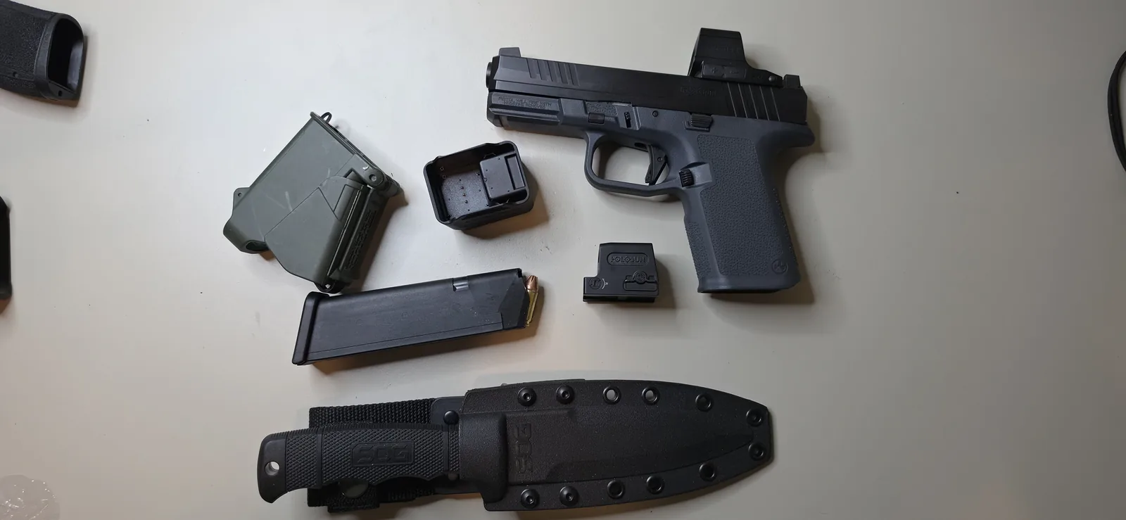 Ruger RXM with a grey Magpul grip frame and an enclosed red dot mounted, beside a spare optic, a magazine, a magazine carrier, and a fixed-blade knife in a kydex sheath